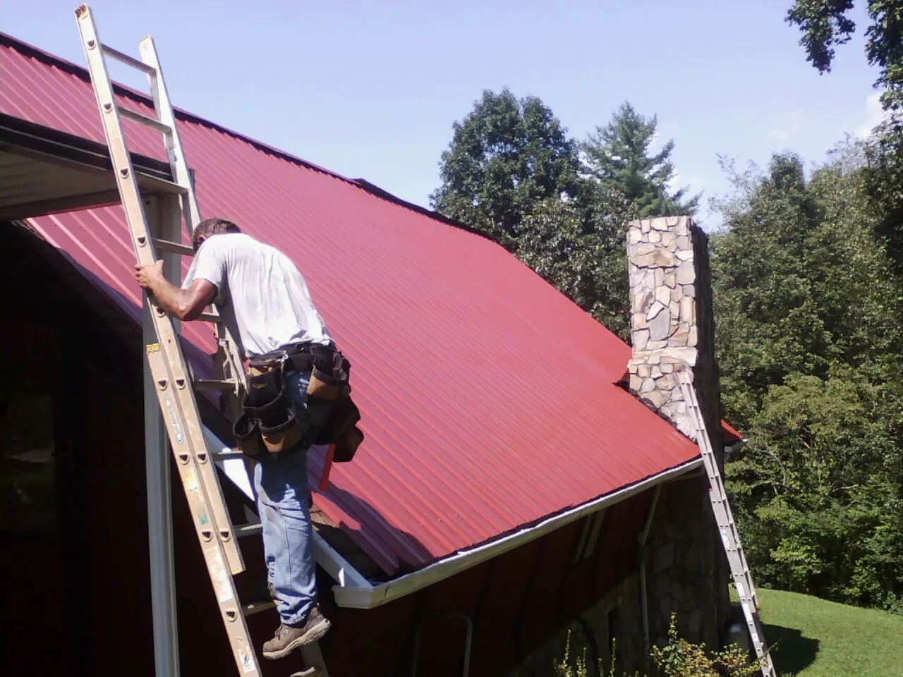Roofer on a ladder beside a red metal roof and stone chimney — Soffit & Fascia Repair by Roofing Brothers in Littleton